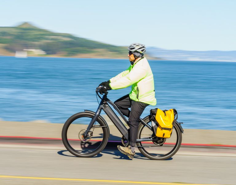 A person in neon gear cycles an e-bike with a yellow pannier on a seaside road.