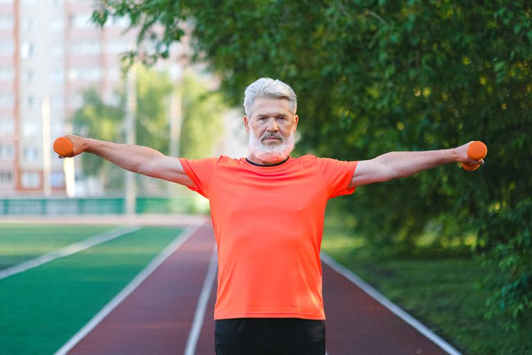 Healthy senior male in activewear exercising with dumbbells on blurred background of stadium and trees