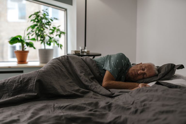 Senior man resting peacefully on a bed inside a room with natural light and plants.