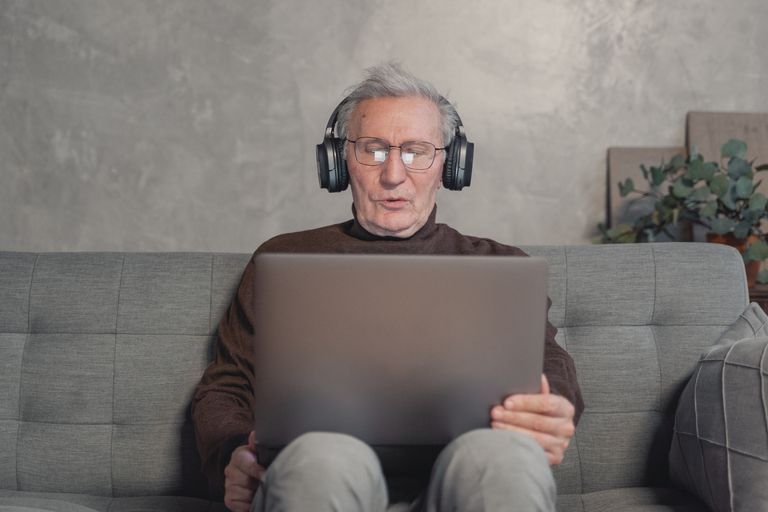 Senior man with headphones working on laptop at home, sitting on a gray couch.