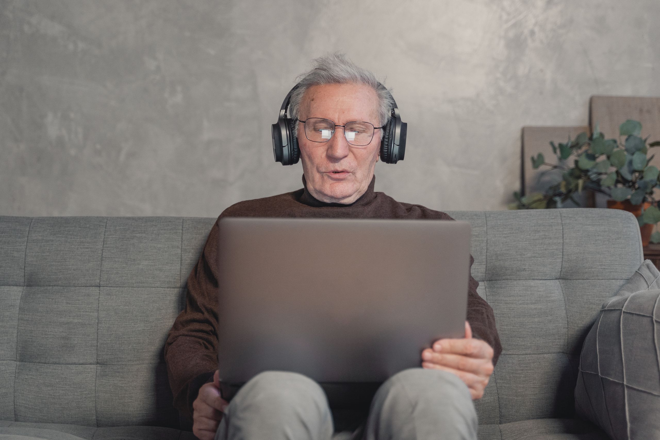 Senior man with headphones working on laptop at home, sitting on a gray couch.