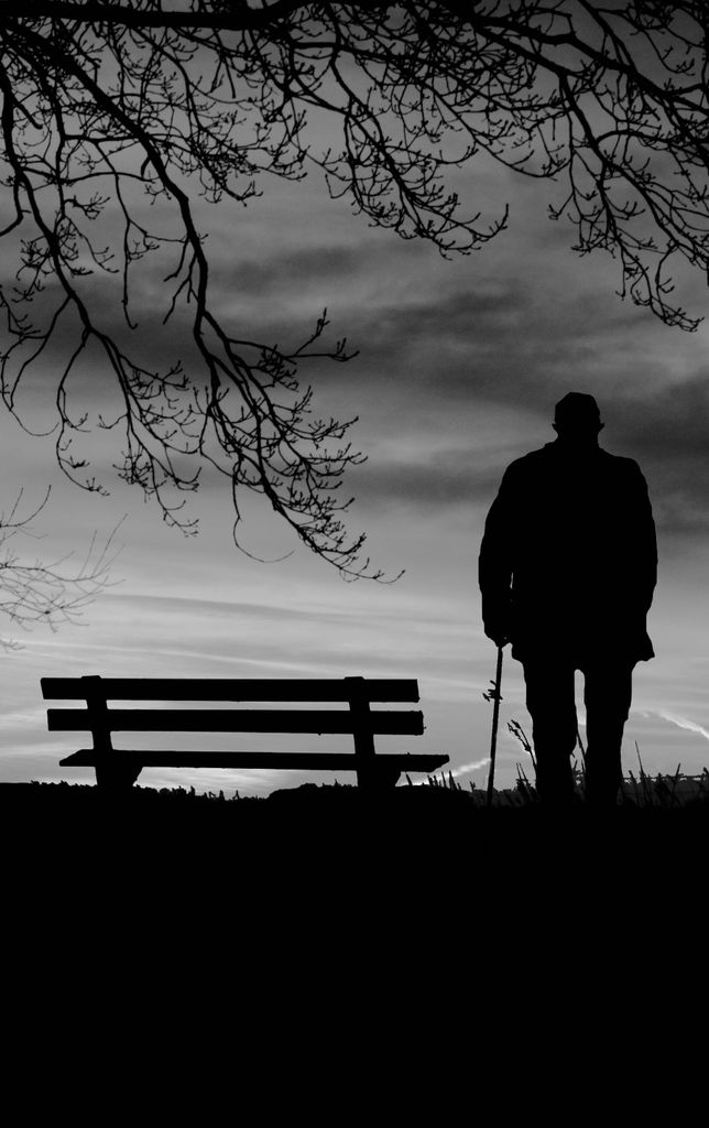 Silhouette of an elderly man with a cane walking near a bench at dusk.