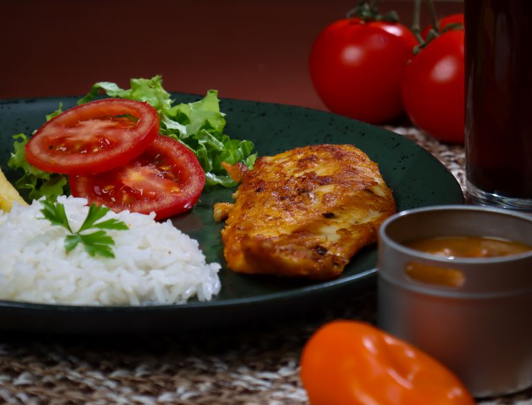 A mouth-watering meal featuring grilled chicken, rice, fresh tomatoes, and salad on a dark plate.