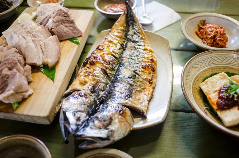 A top view of a platter with sliced pork, grilled mackerel, kimchi, and other Korean dishes on a wooden table.