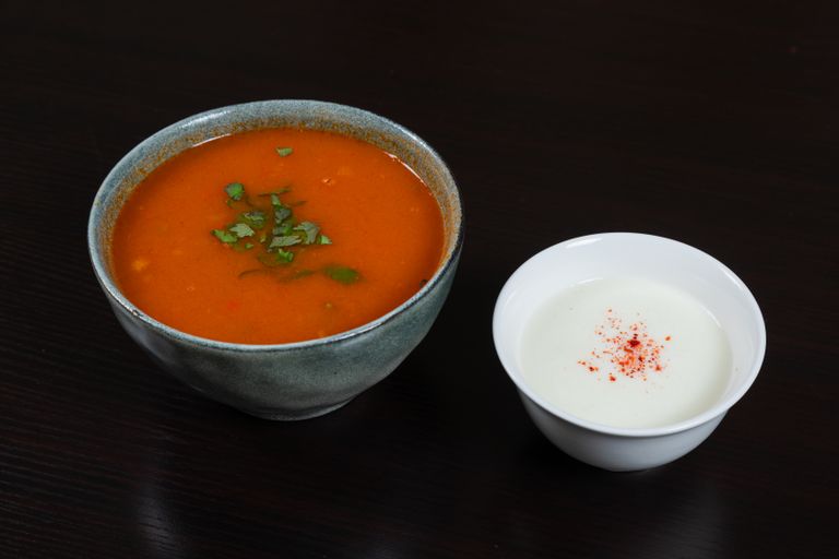 Bowl of tomato soup garnished with herbs served alongside a creamy dip on a dark surface.