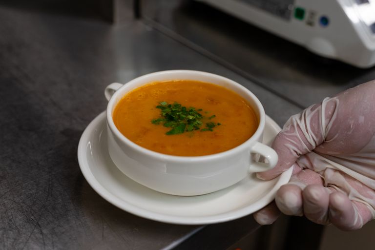 Close-up of a chef's hand holding a ceramic bowl of tomato soup garnished with herbs.