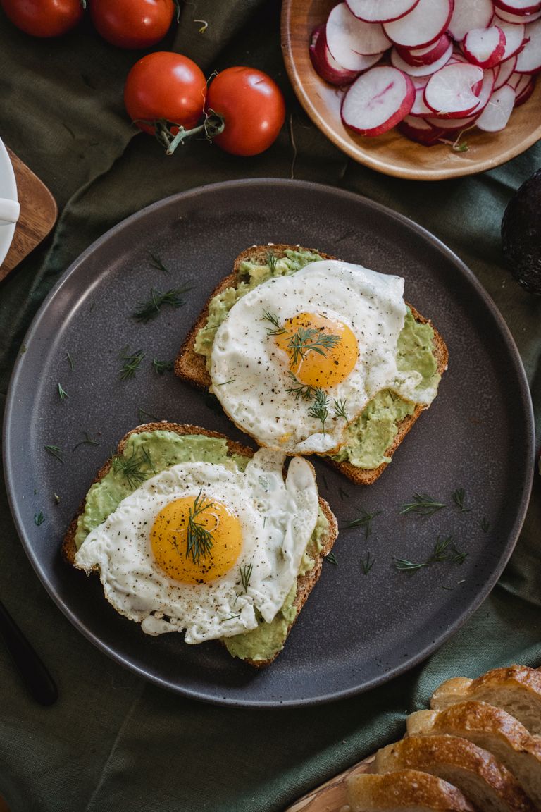 Delicious avocado toast topped with sunny side up eggs, served with fresh radishes and tomatoes.