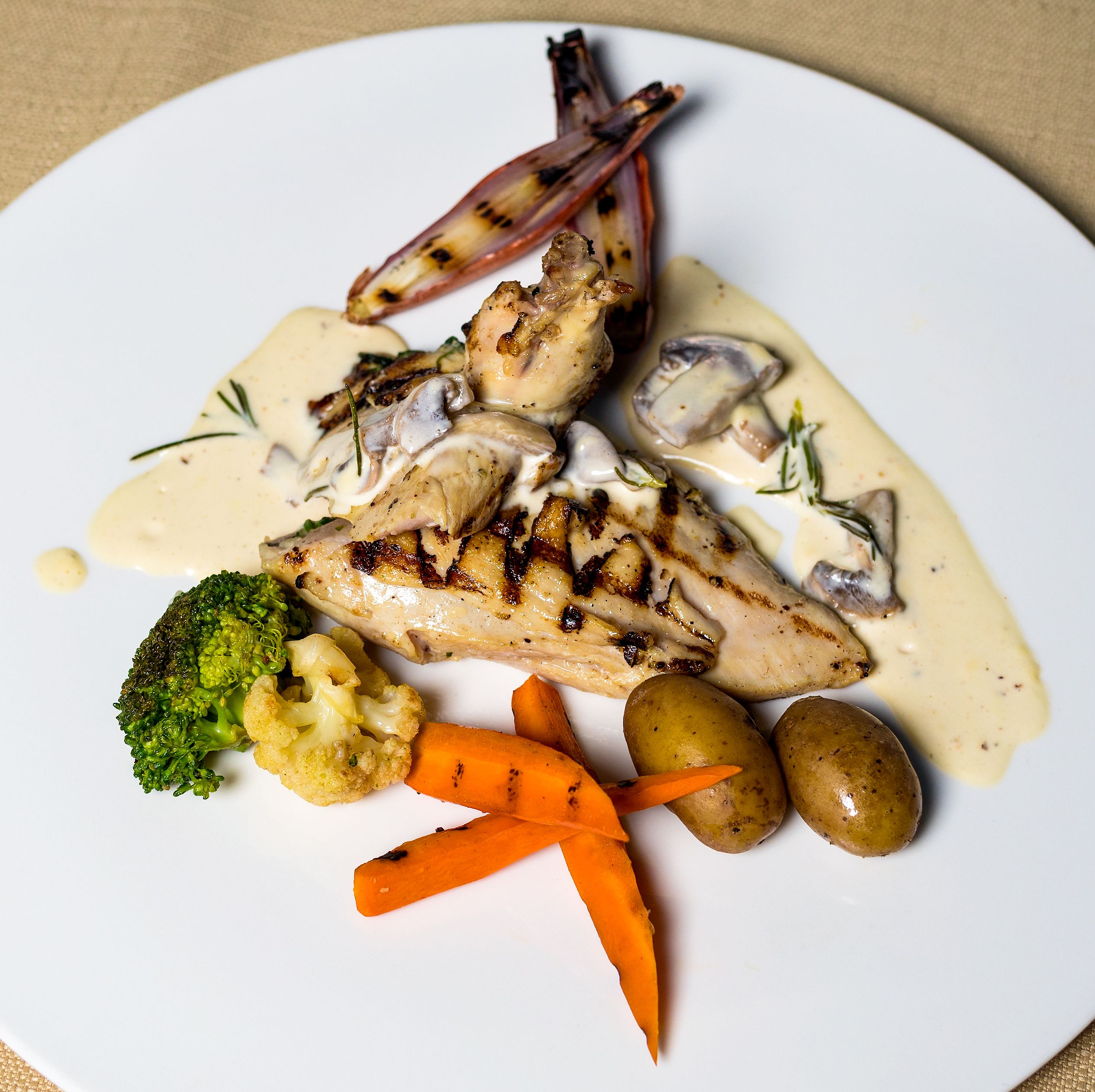 Delicious grilled chicken breast with creamy sauce, vegetables, and potatoes on a plate.