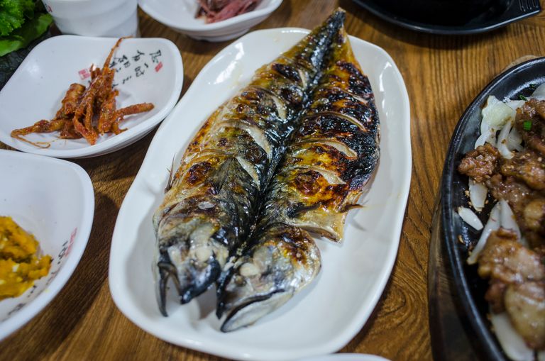 Delicious grilled mackerel on a white plate surrounded by Korean side dishes, showcasing authentic cuisine.