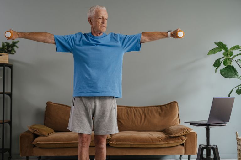 Elderly man in blue shirt exercising with dumbbells at home, enhancing fitness and wellness.