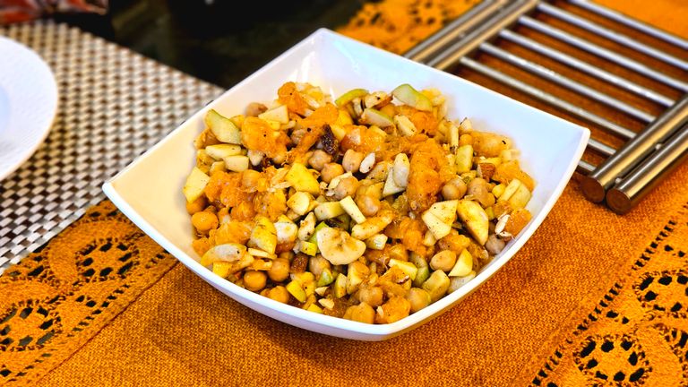 Freshly tossed chickpea and fruit salad with oranges and apples in a white bowl on a patterned tablecloth.