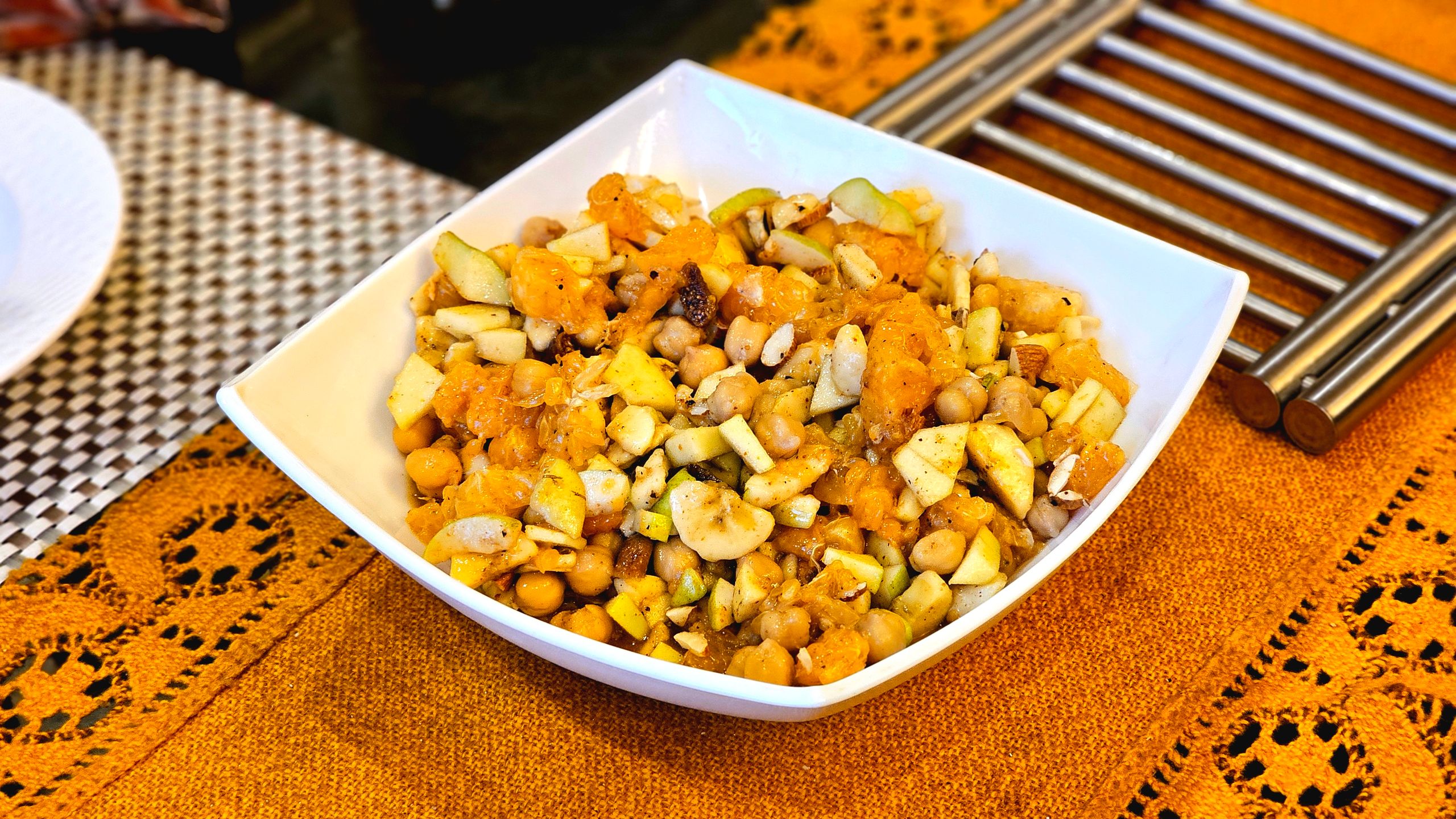Freshly tossed chickpea and fruit salad with oranges and apples in a white bowl on a patterned tablecloth.