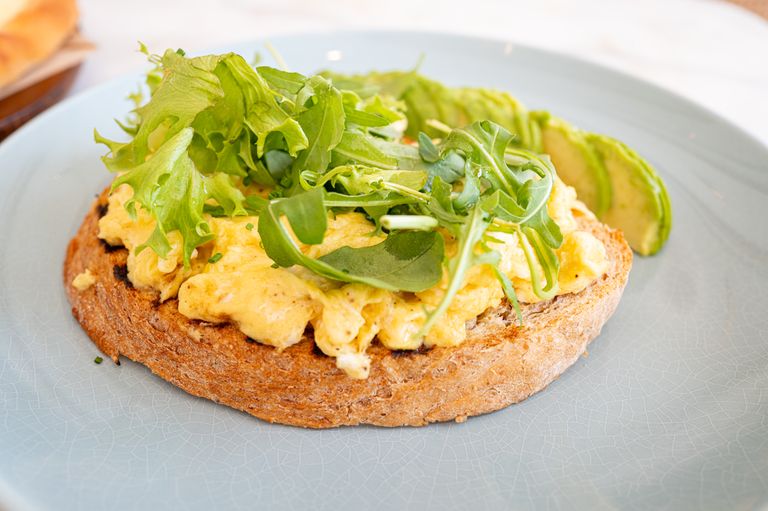Healthy breakfast featuring scrambled eggs, avocado, and greens on toast on a blue plate.