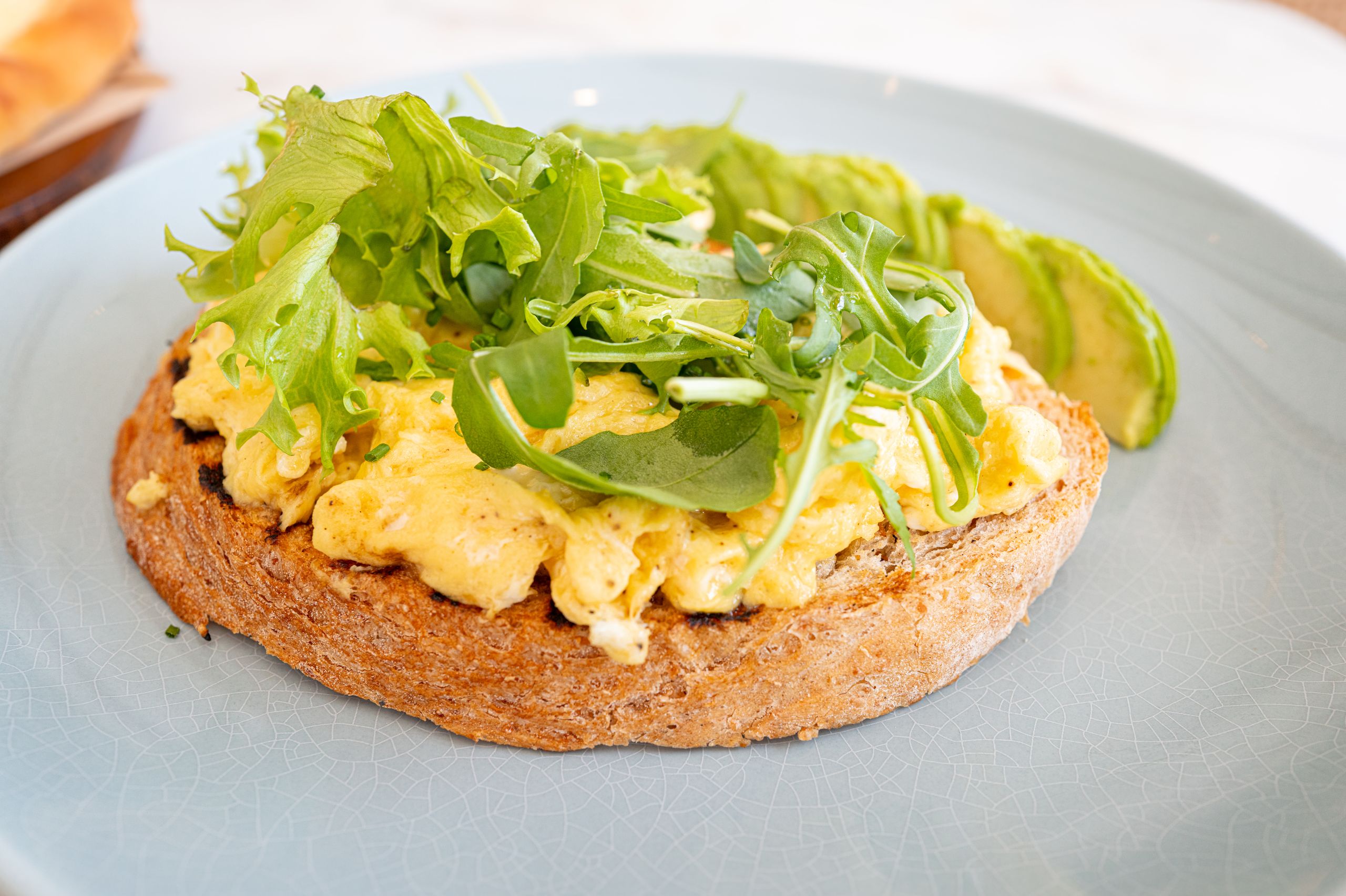 Healthy breakfast featuring scrambled eggs, avocado, and greens on toast on a blue plate.