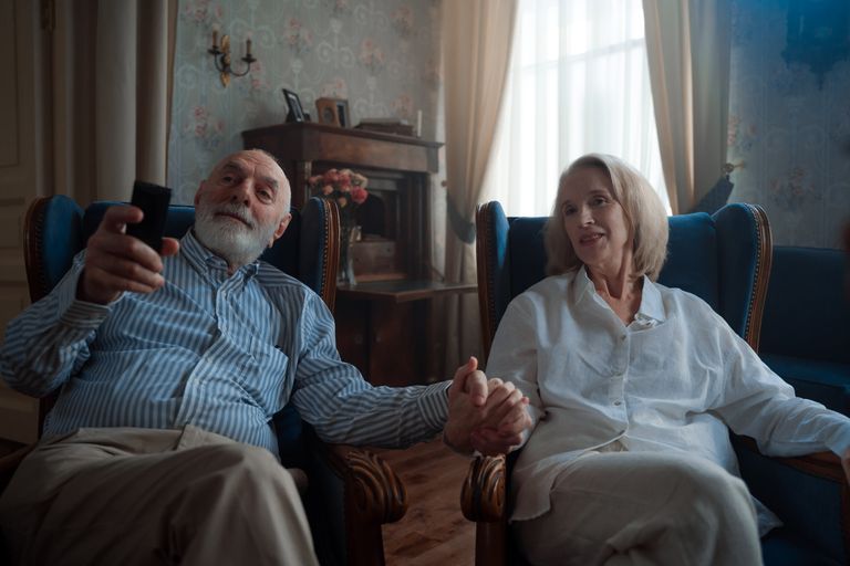 Senior couple sitting together, holding hands and watching TV in a cozy living room.