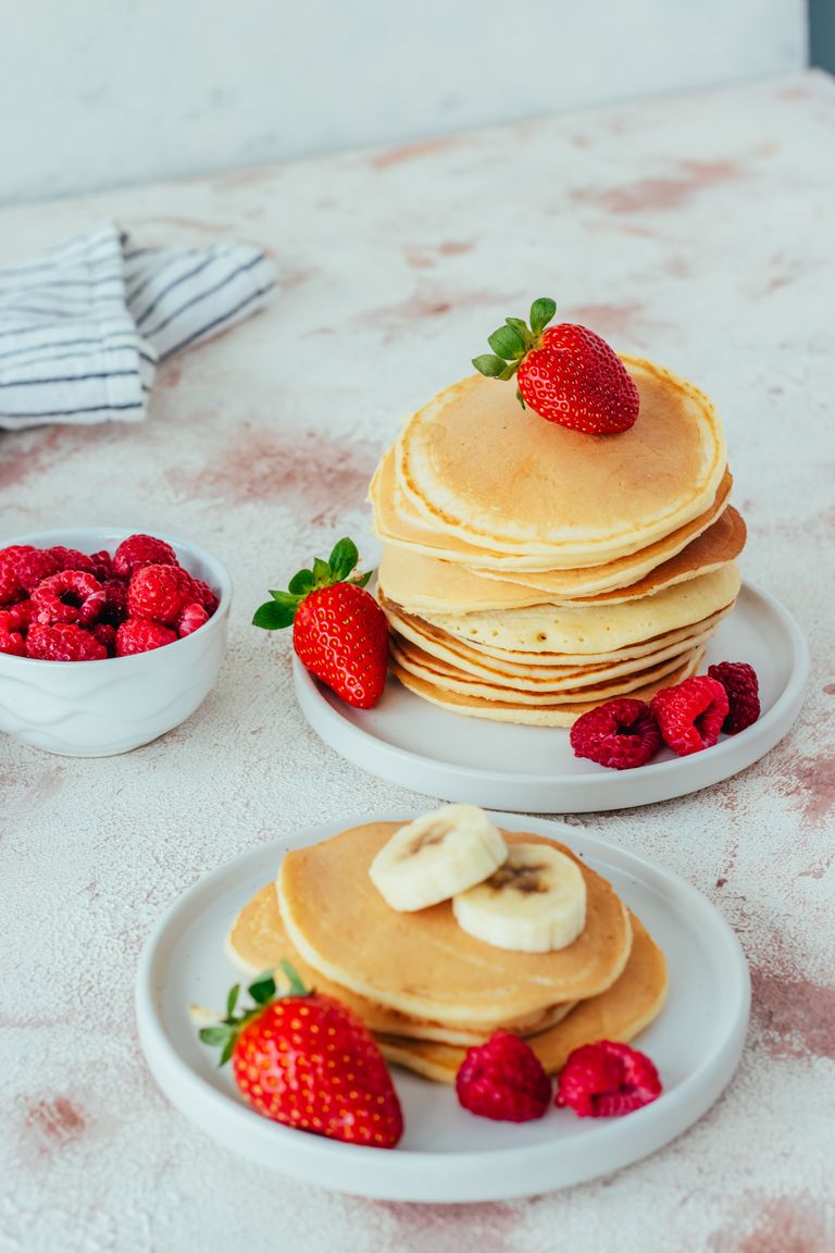 Stack of pancakes topped with strawberries and bananas, accompanied by bowls of fresh raspberries.