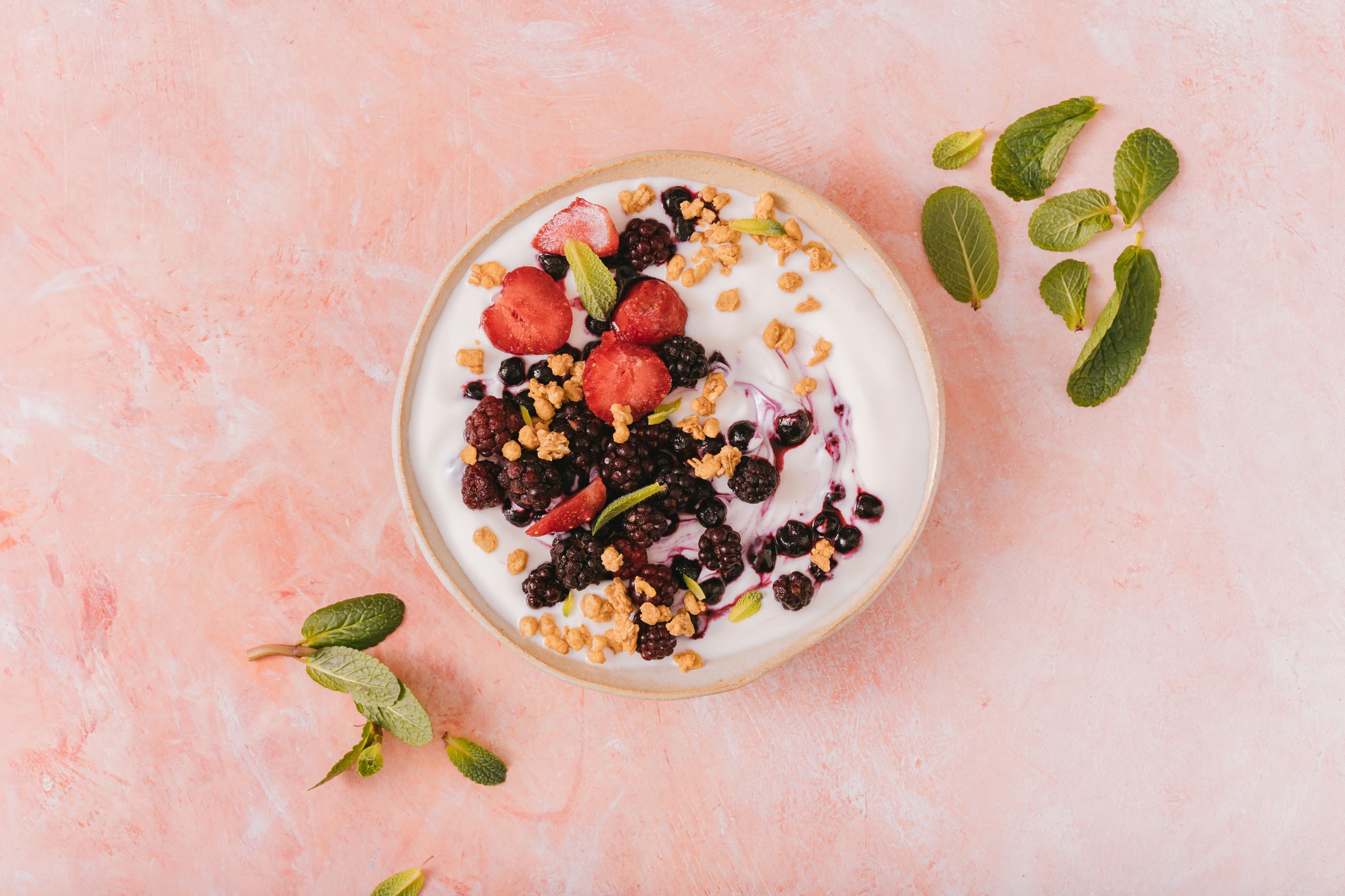Top view of a vibrant yogurt bowl with mixed berries, granola, and mint leaves on a pink background.