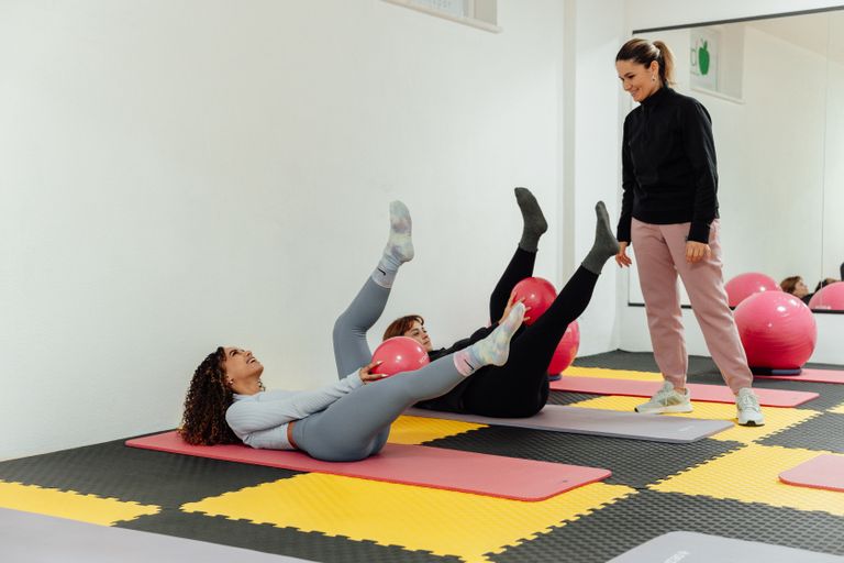 Two women exercising with pink balls on yoga mats under trainer supervision in a Yalova gym.